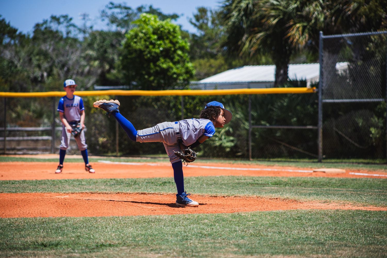 Baseball pitcher in mid-windup delivers pitch from dirt mound on sunny baseball field.