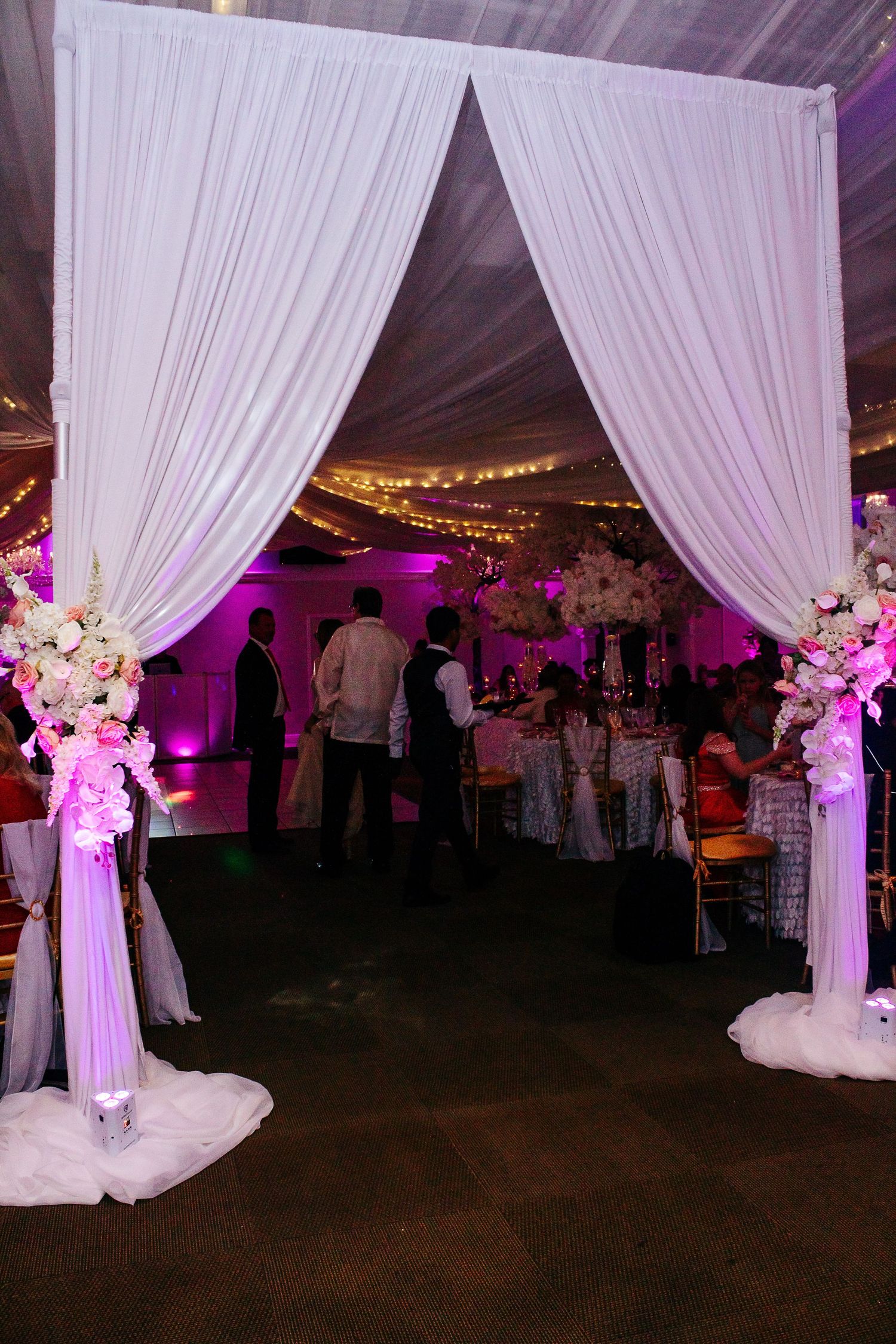 Elegant wedding venue entrance with white draping, pink uplighting and floral arrangements on pedestals.