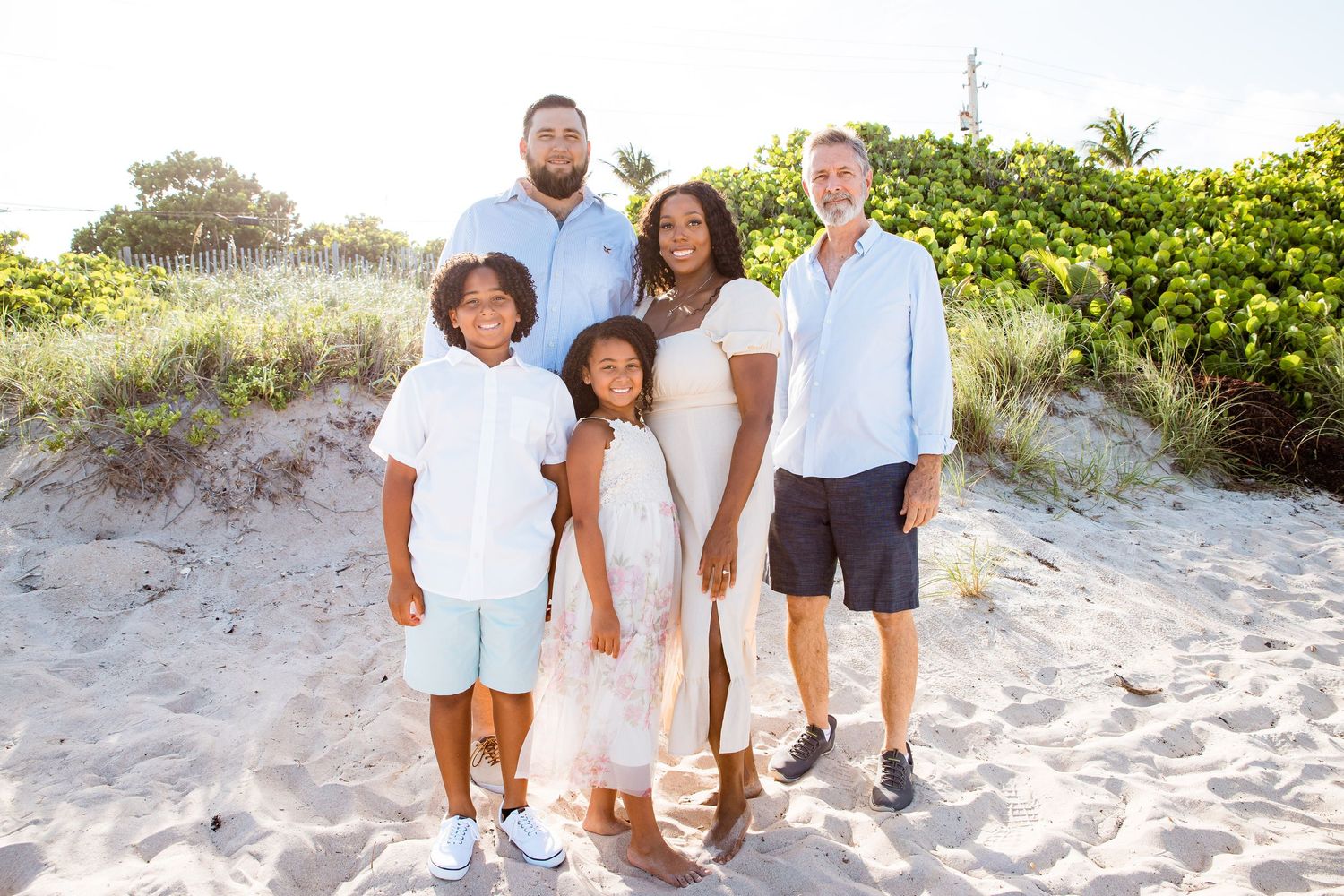 A family stands together smiling on a sandy beach with green foliage in the background during golden hour.