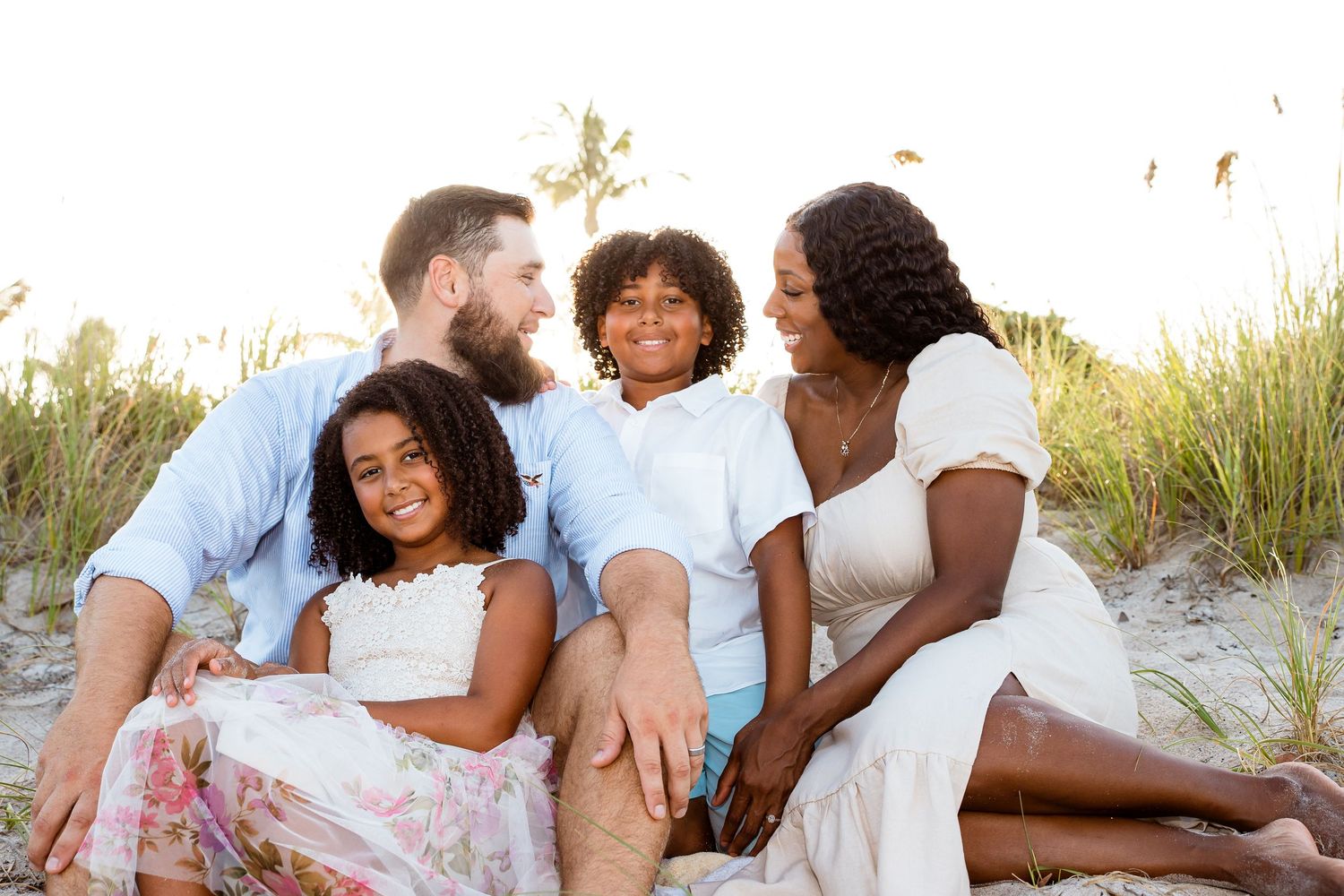 A family sits together smiling at sunset on a beach with tall grass in the background.