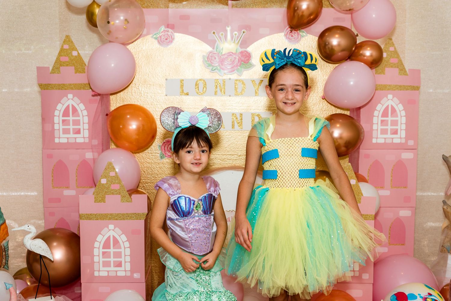 Pink and gold balloon decorations create a princess castle backdrop with two children in sparkly tulle dresses.