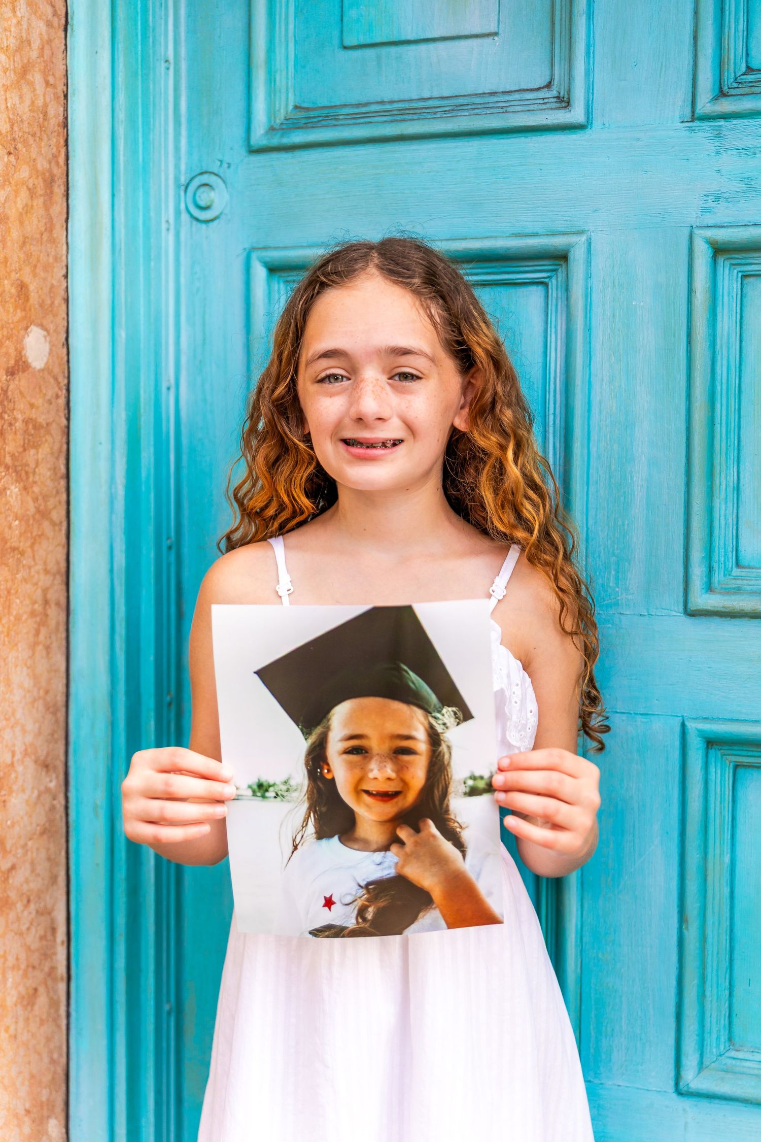 Young girl in white dress holds graduation photo portrait in front of teal blue door.