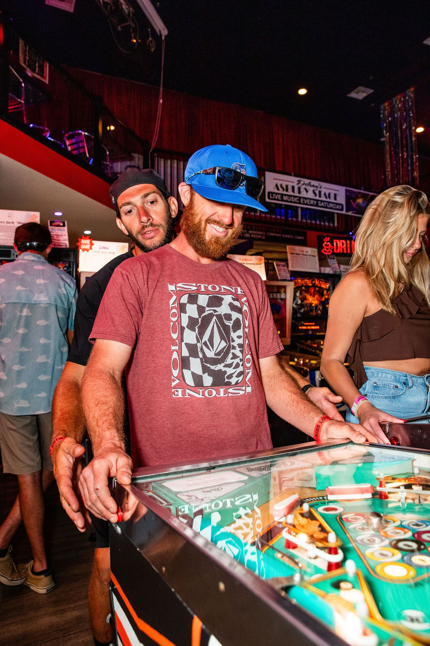 Two men playing pinball at a busy arcade bar, one wearing a blue cap and Volcom t-shirt.