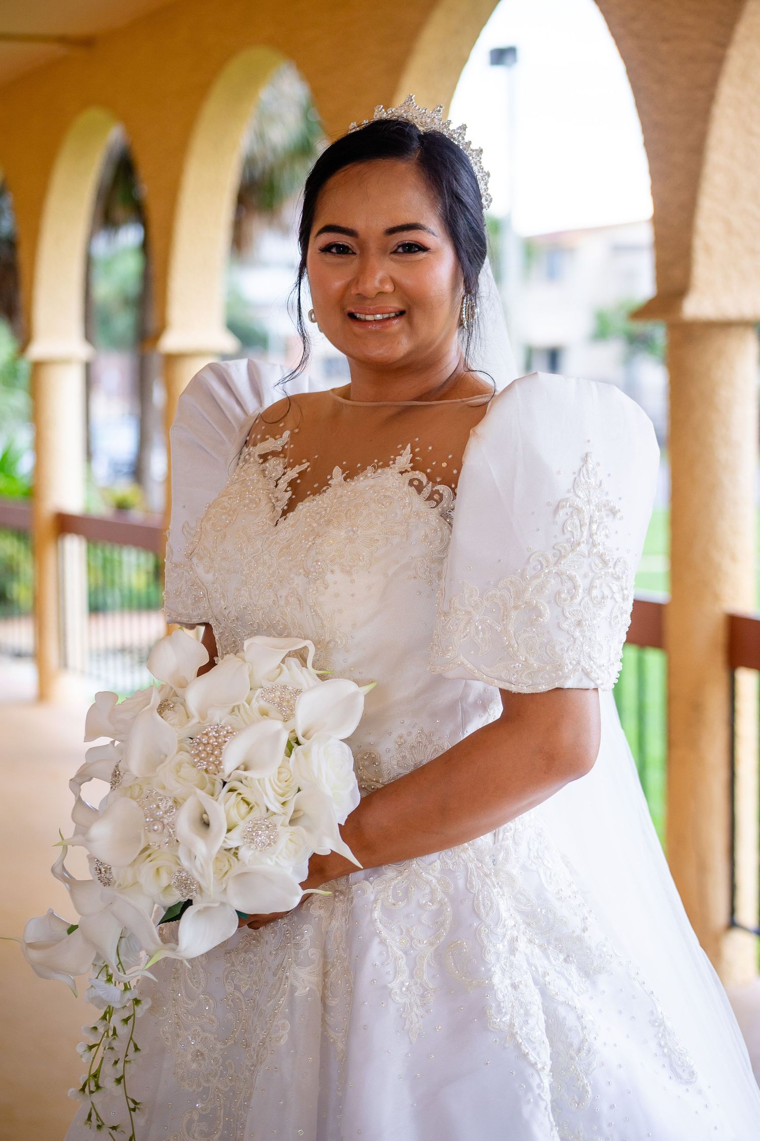 Bride in lace wedding dress with puff sleeves poses with white orchid bouquet under arches.