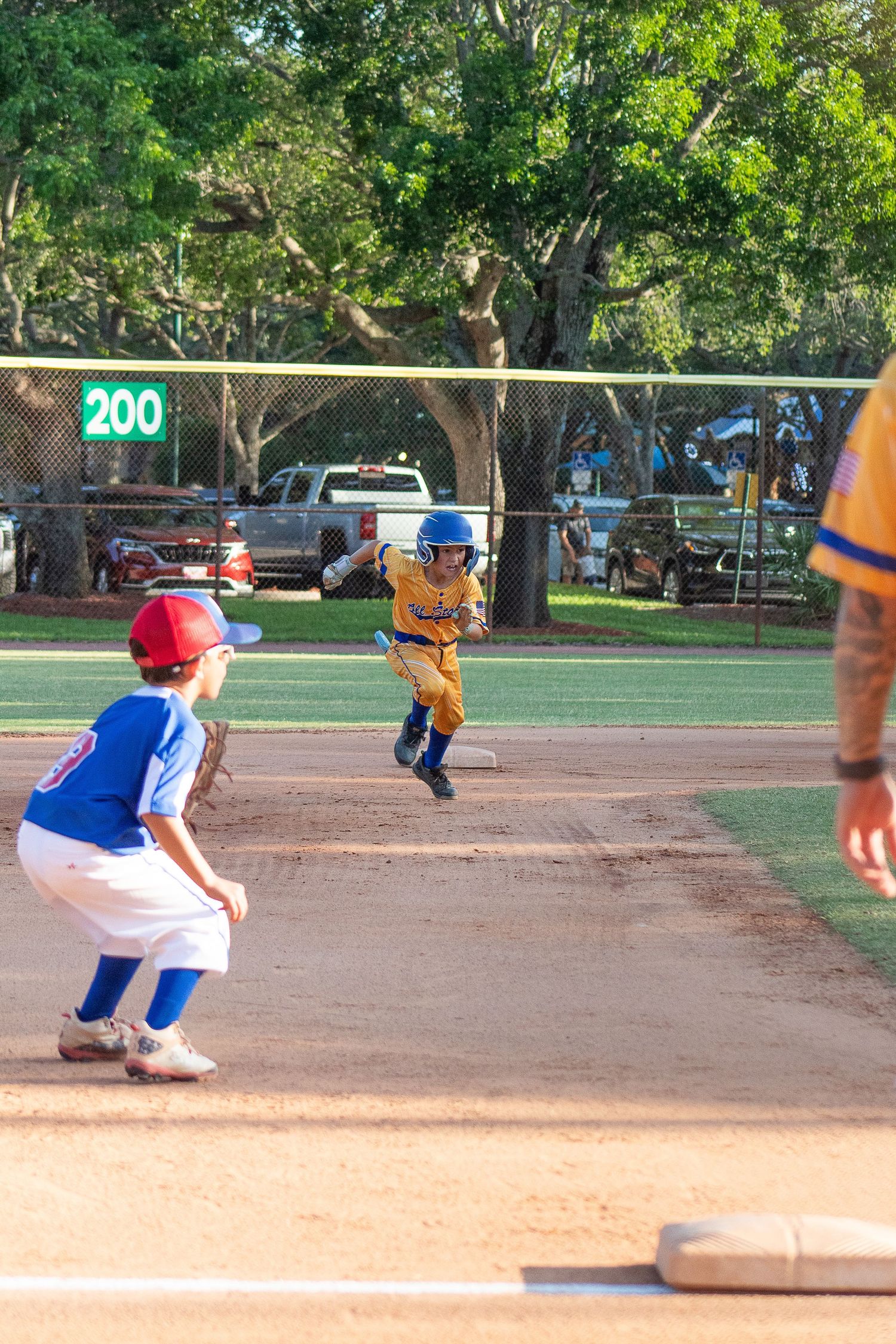 Baseball players in action during youth league game with scoreboard visible in background.