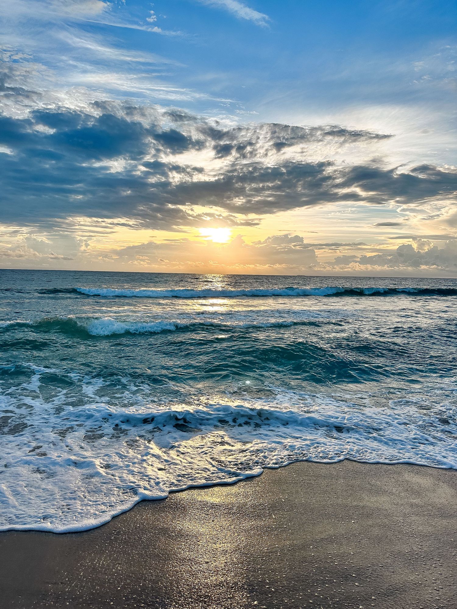 Dramatic sunset over ocean waves crashing on dark sandy beach with vibrant clouds in the sky.