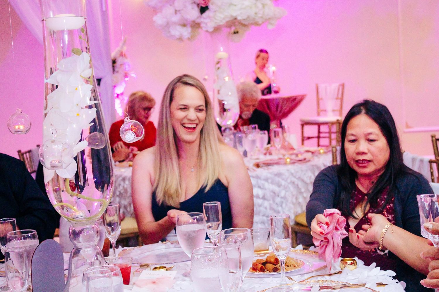 Wedding reception guests seated at decorated tables with pink ambient lighting.