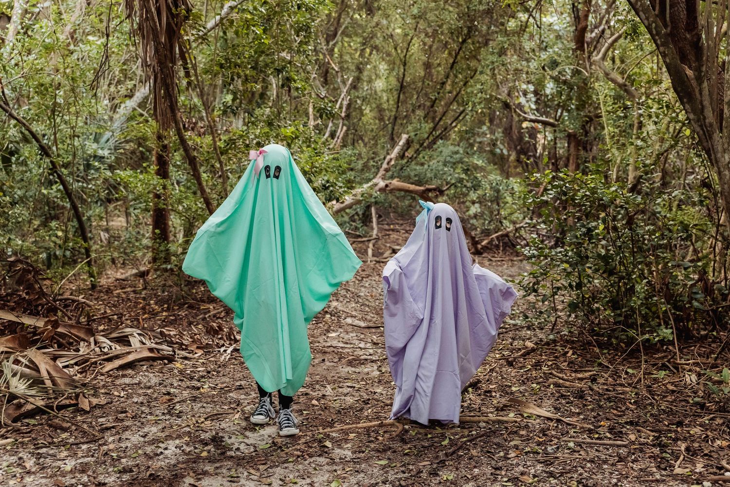 Two figures wearing ghost-like costumes in green and purple walk through a wooded forest path.