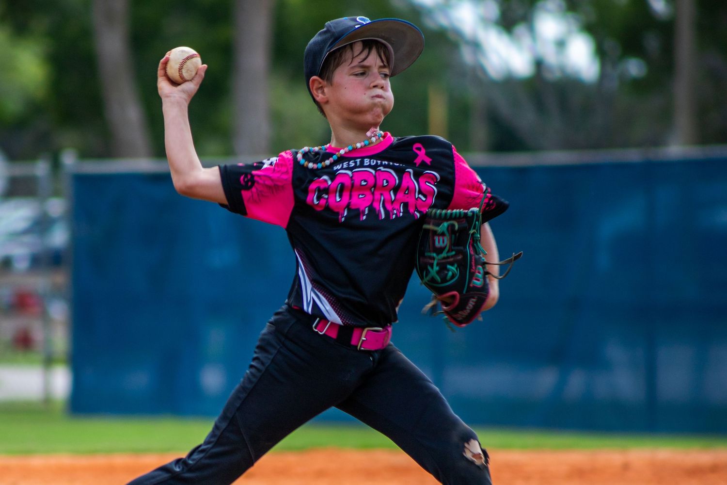 Baseball pitcher in black and pink uniform winds up to throw pitch during game on outdoor field.