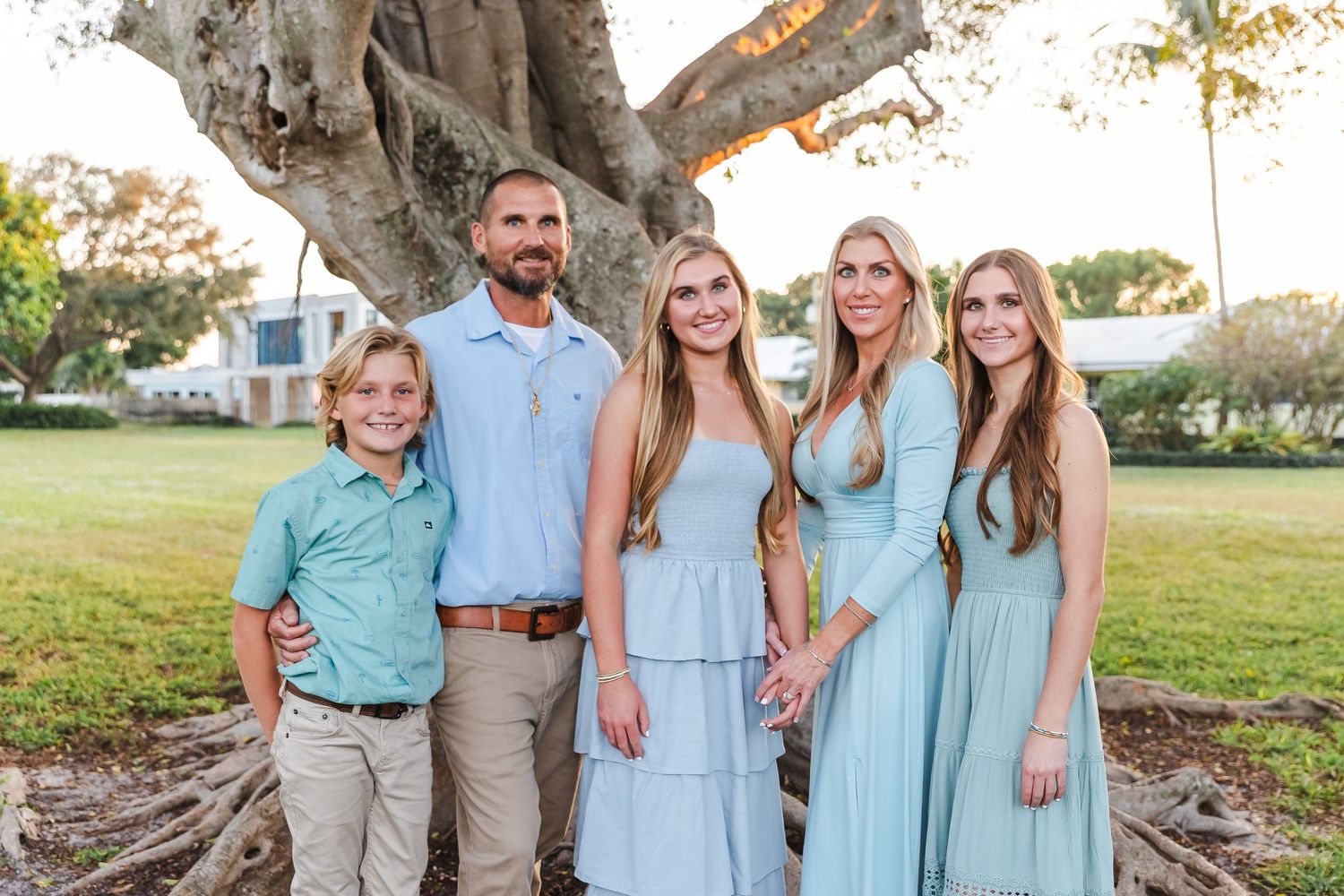 A group in coordinated light blue and beige outfits poses together under a large tree in a park setting.