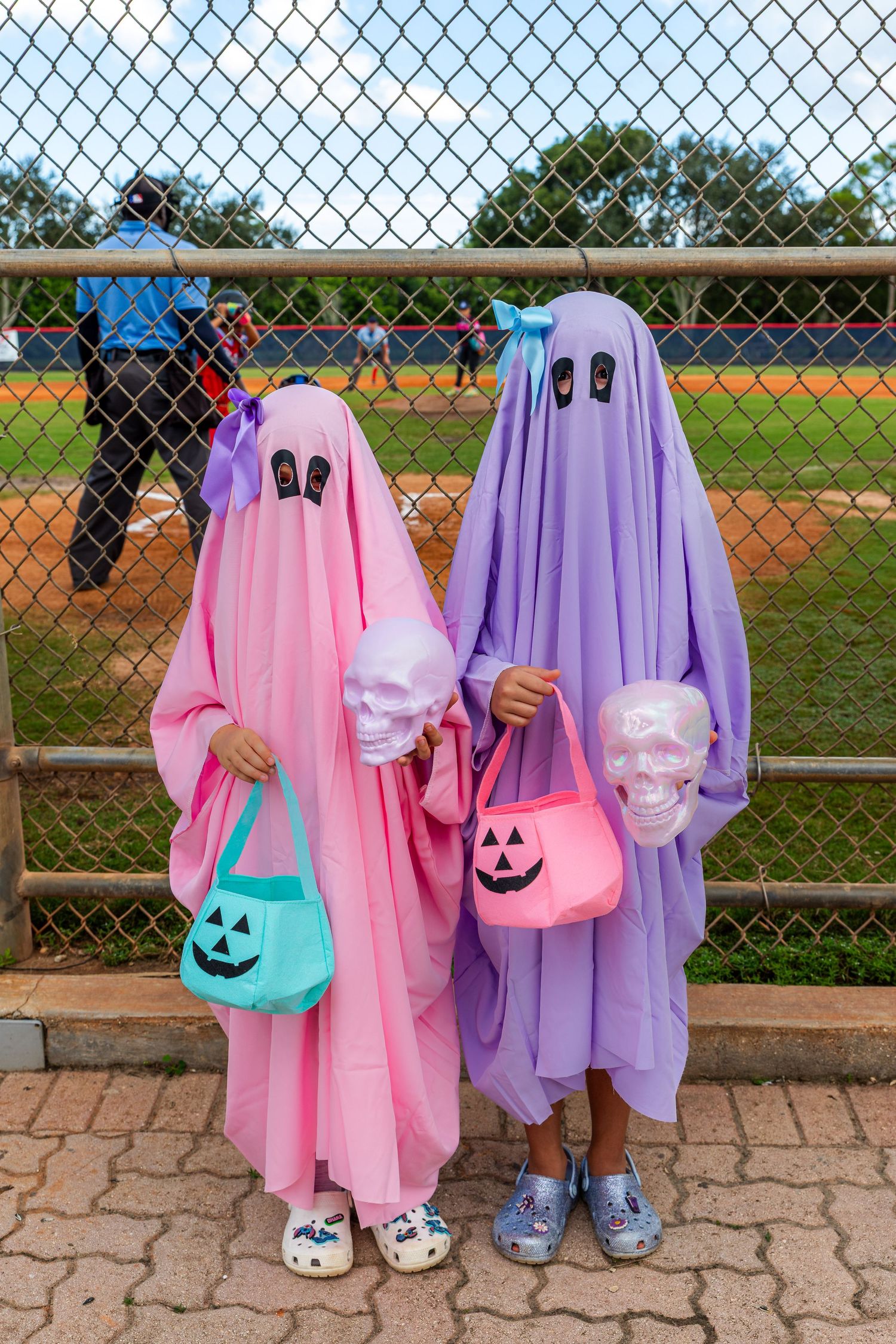 Two children wearing pink and purple ghost costumes with smiling faces stand together at a baseball field.