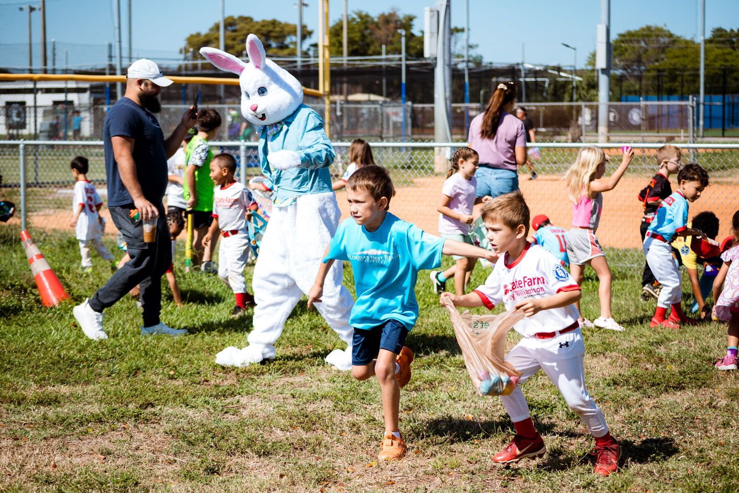 Children run excitedly on a grassy field during an Easter egg hunt with a bunny mascot at a community event.