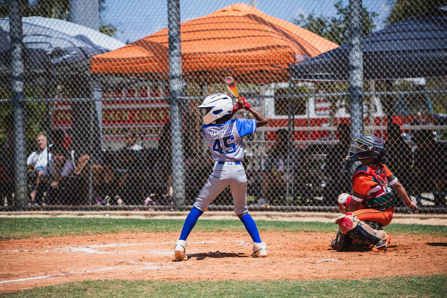 Baseball player at bat with a catcher crouching behind home plate on a dirt diamond with orange canopies visible.