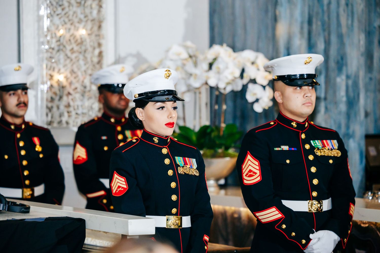 Military service members in dress uniforms lined up at a formal event with decorative lighting.