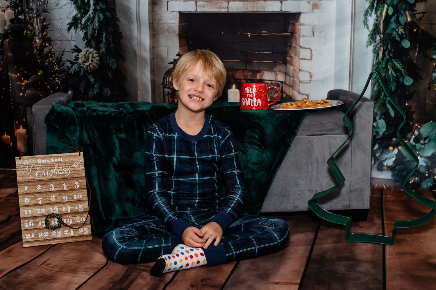 Child in plaid pajamas sits cross-legged by a fireplace with holiday decorations.