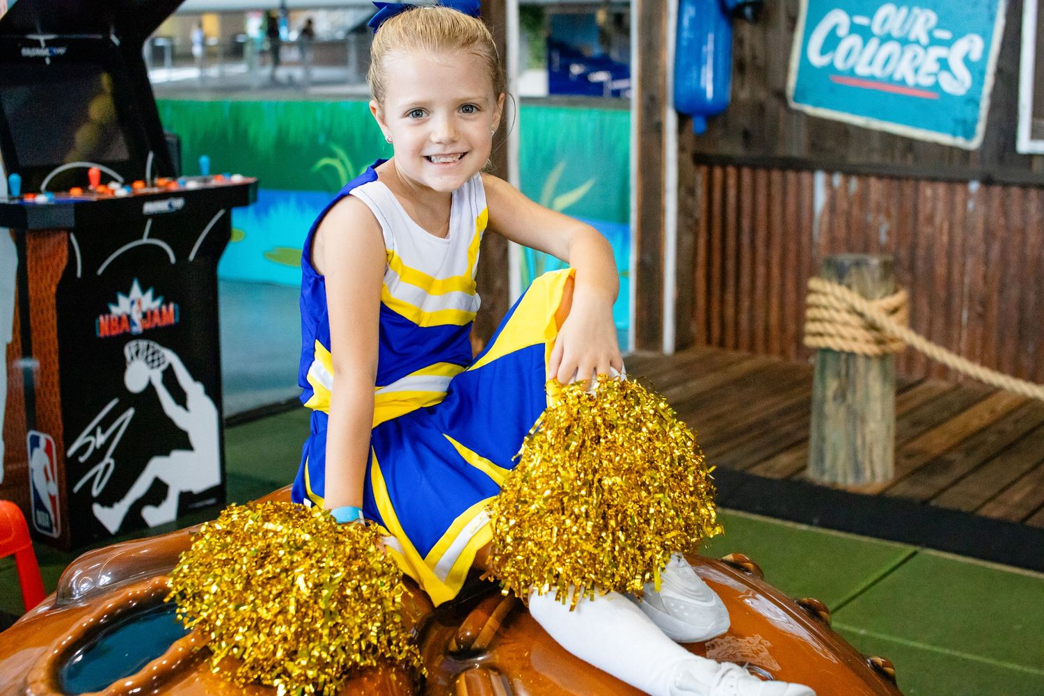Young cheerleader sitting with gold pom poms in rustic sports-themed setting.
