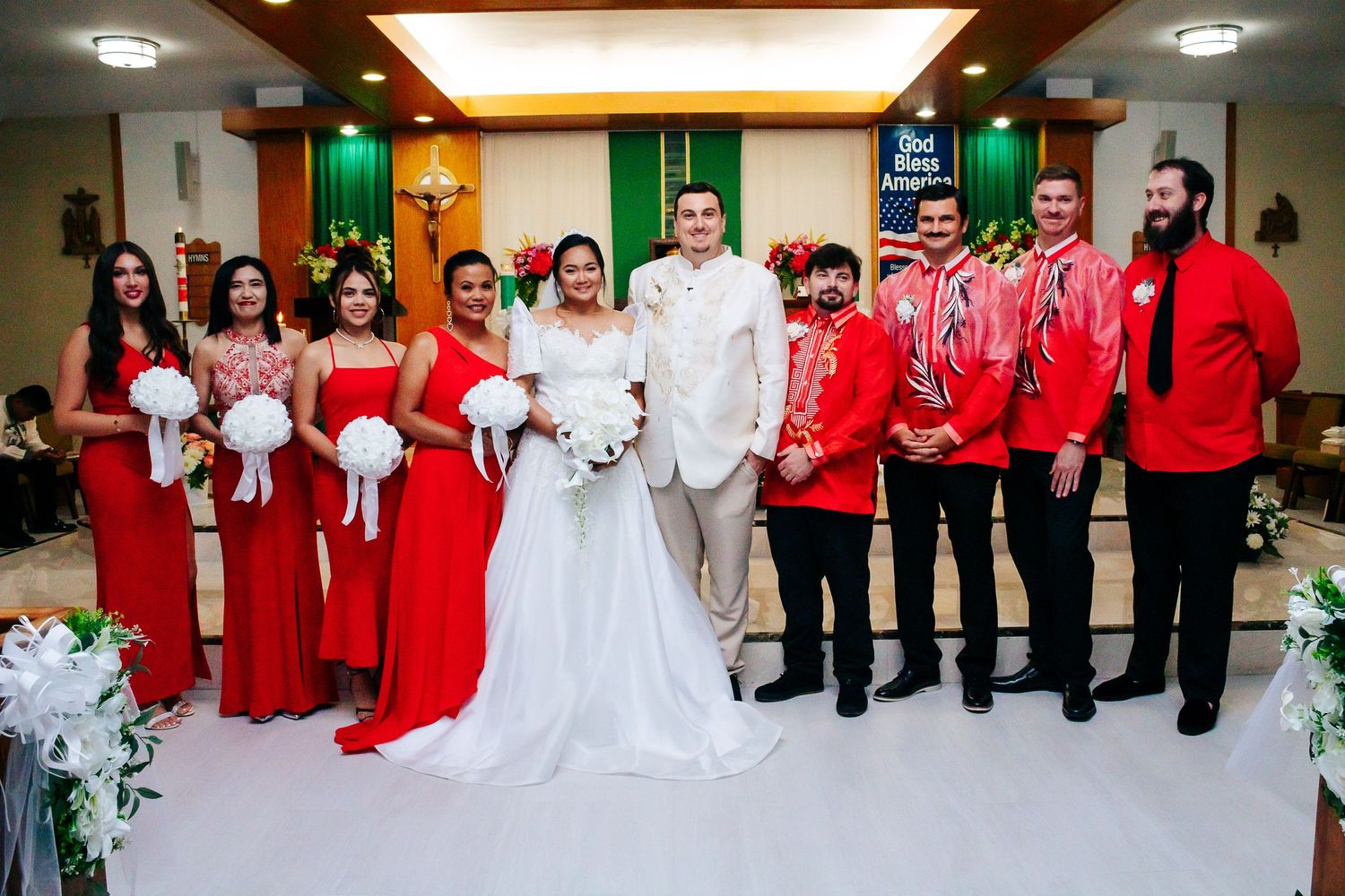 Large wedding party in red and white attire gathered for formal photo at church.