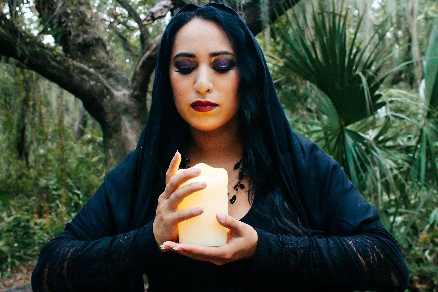 A figure in dark clothing holds a glowing candle in a mystical forest setting.