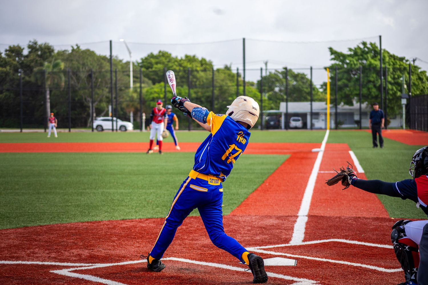 Young baseball player in blue and yellow uniform swings bat at home plate on a red turf field.
