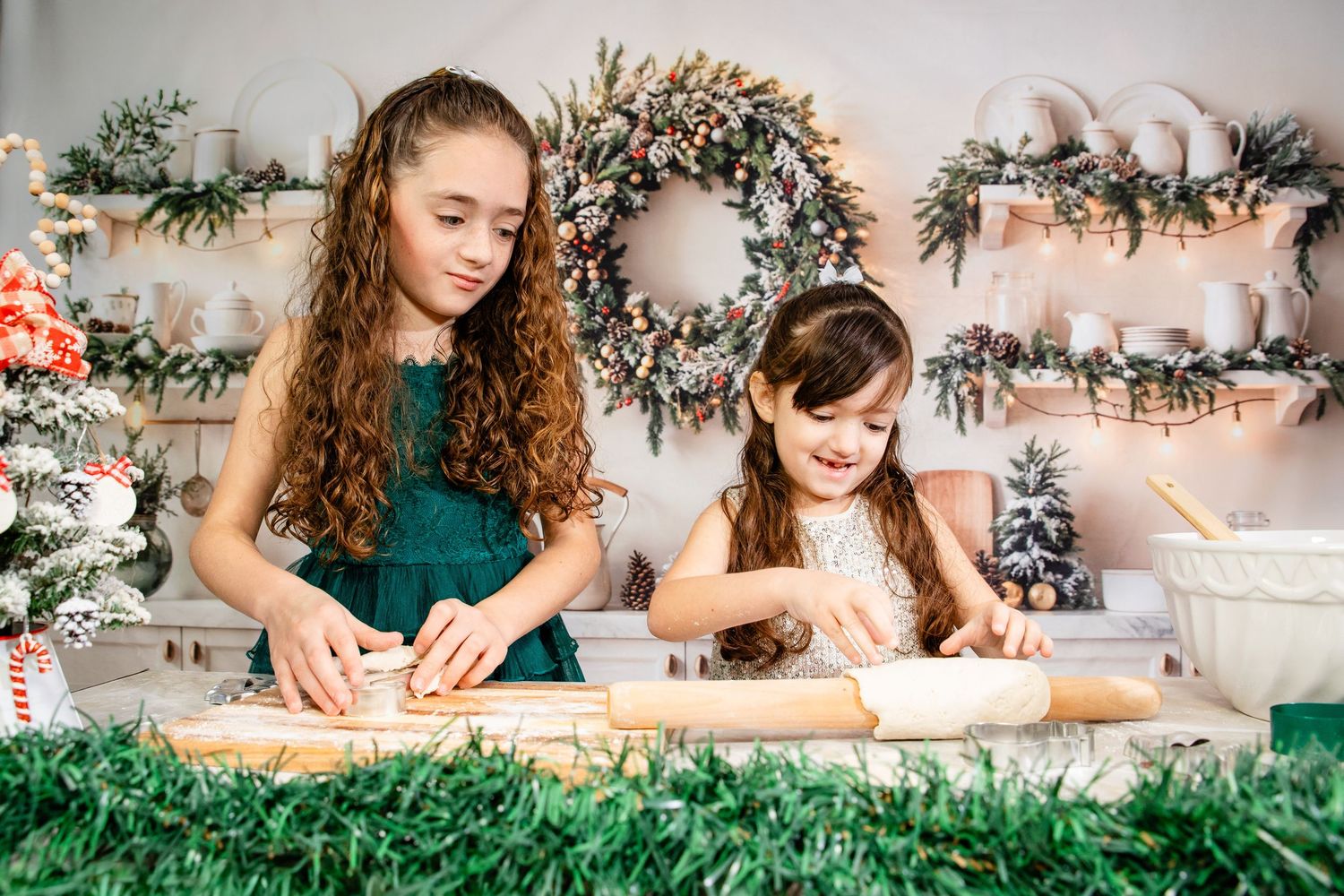 Hands rolling out dough on a wooden surface with holiday decorations and greenery in the background.