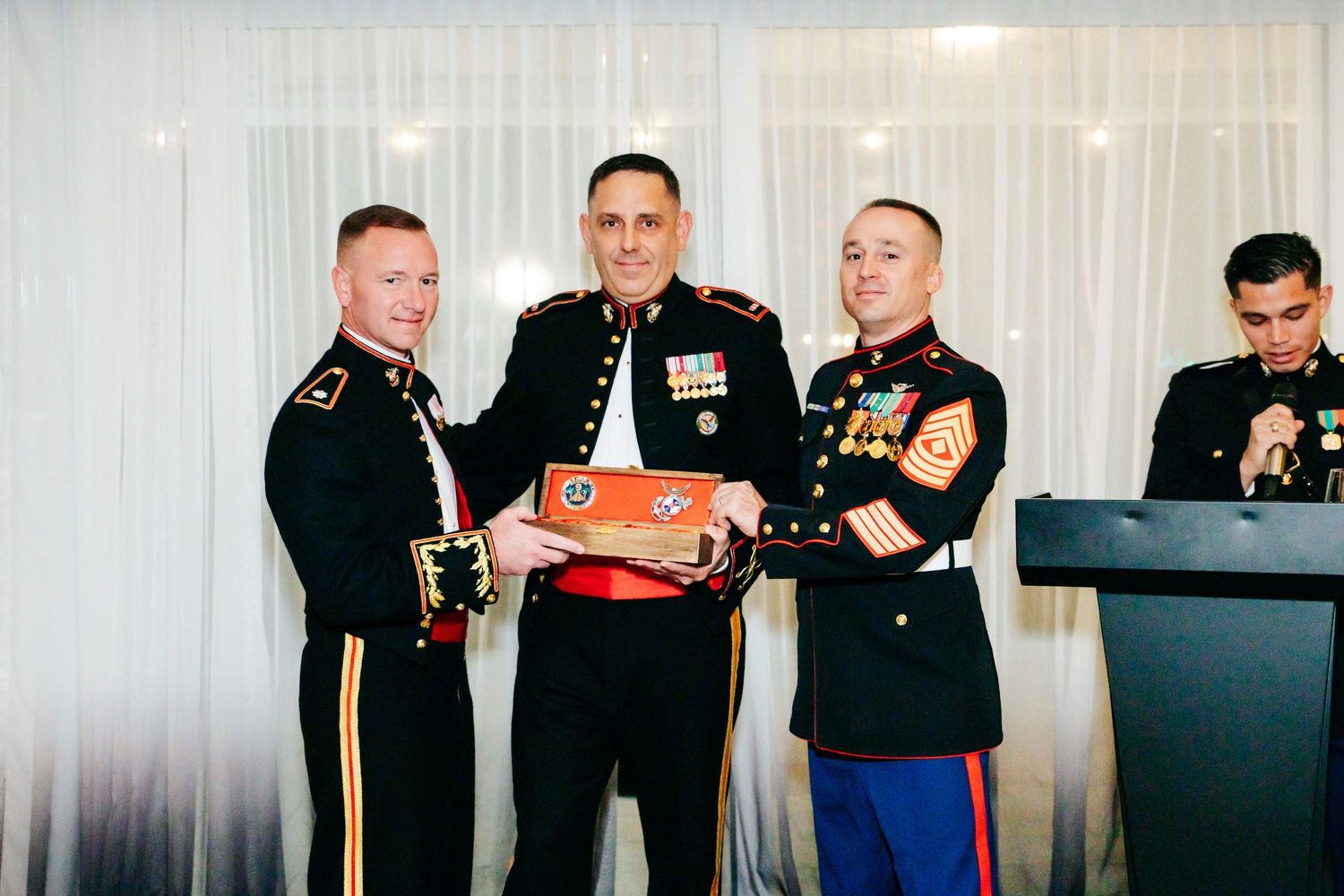 Marines in dress uniforms socialize at formal military event with white backdrop.