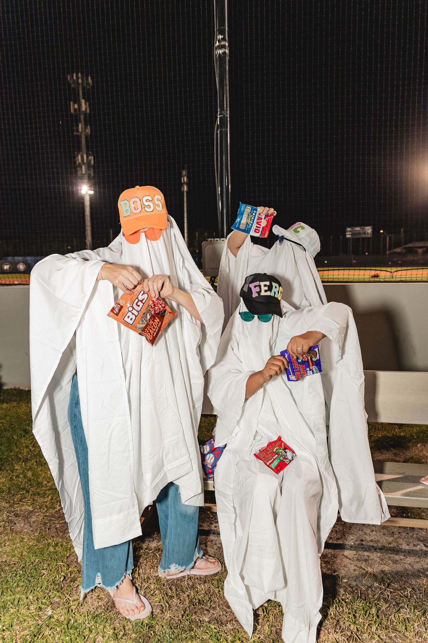 People wearing white ghost costumes with orange and colorful accessories pose at night in a baseball dugout.