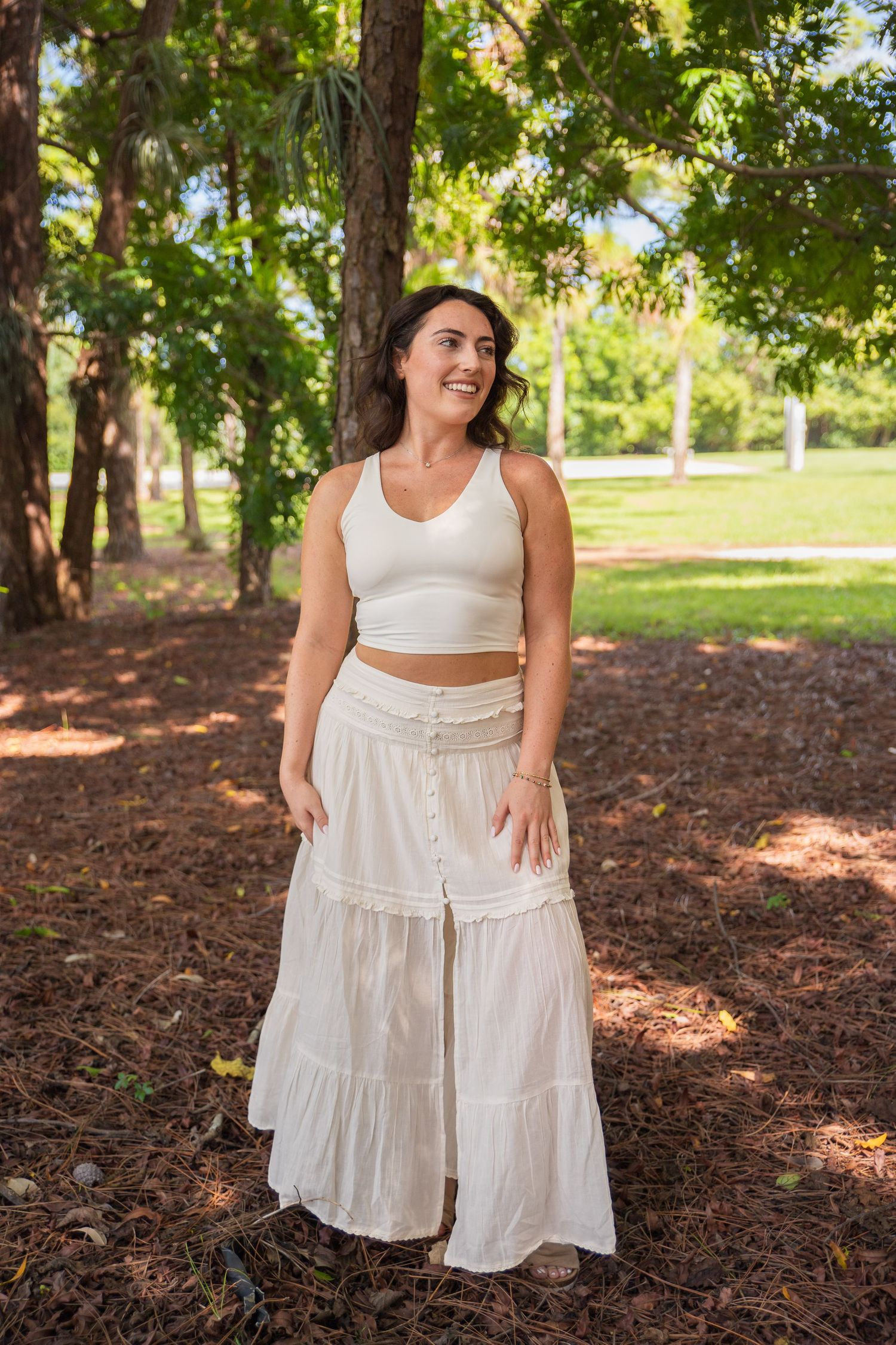 A person wearing a white crop top and flowing maxi skirt stands in a wooded park on a sunny day.