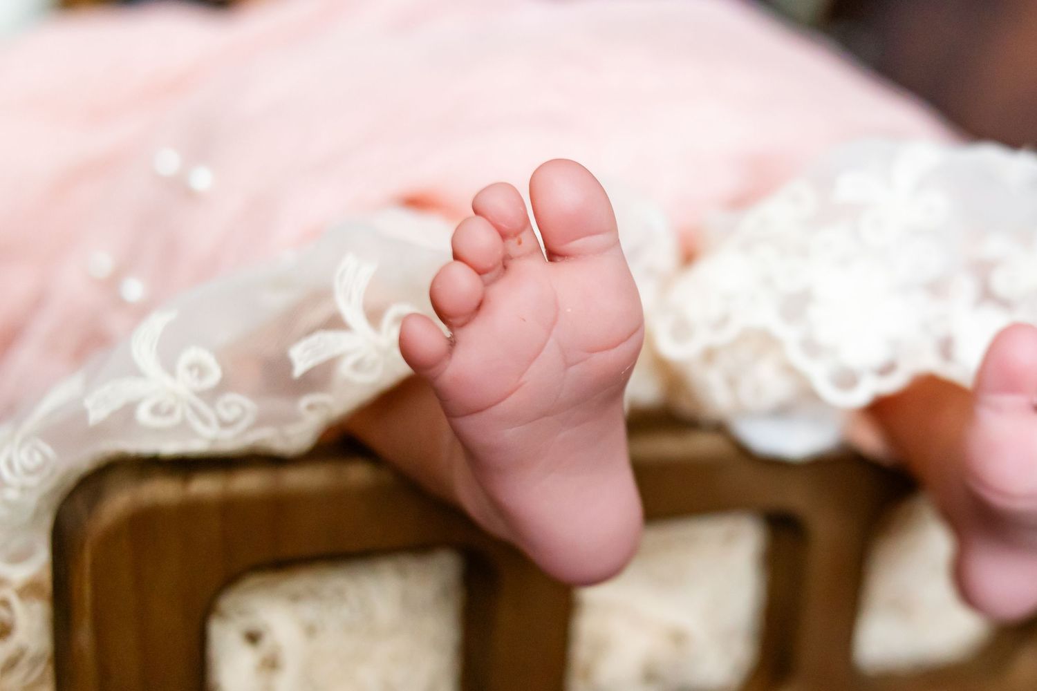 Close-up of tiny newborn baby feet peeking out from a delicate pink lace dress while resting in a wooden crib.