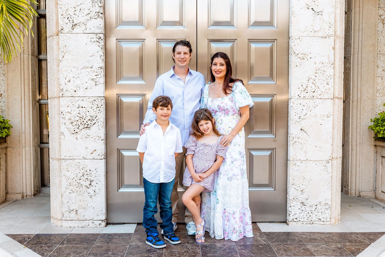 Family of four posing in front of elegant large wooden doors, parents and two children smiling in coordinated outfits.