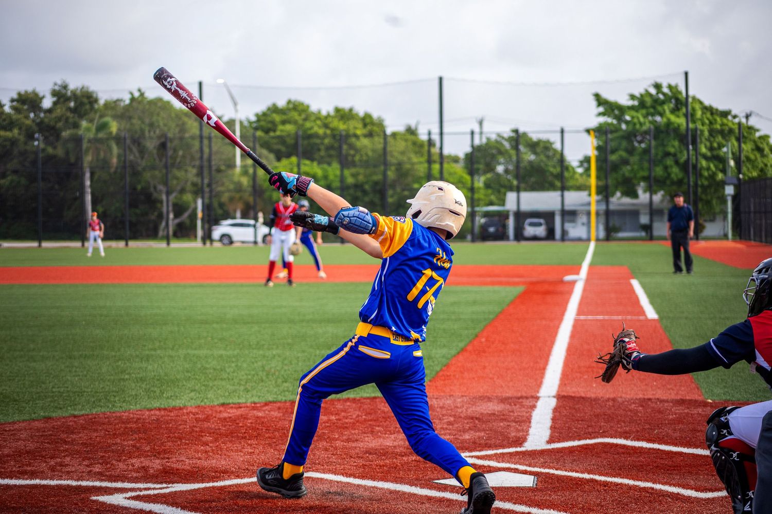 Young baseball player in blue and yellow uniform swinging bat during a game on a red dirt field.