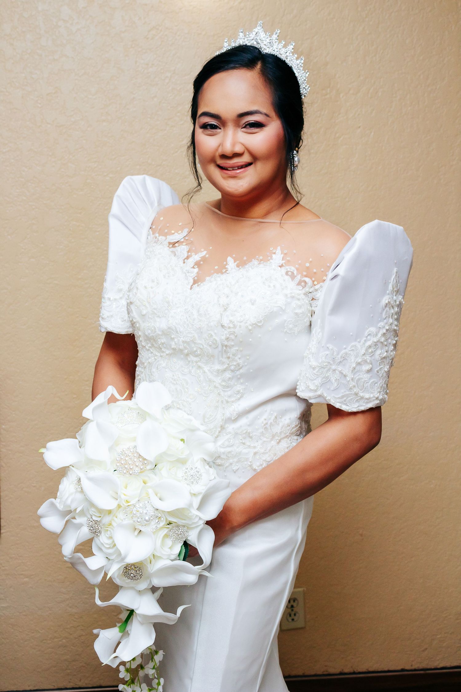 Beautiful bride in white wedding gown holding elegant white orchid bouquet against beige background.