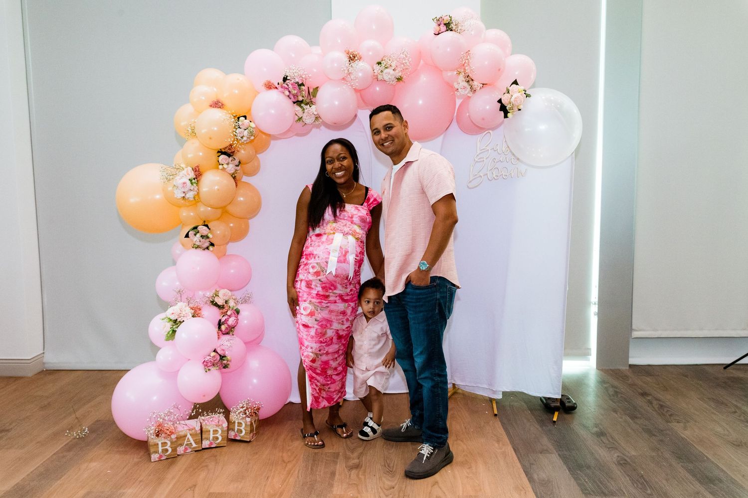 A couple poses in front of a decorative pink and peach balloon arch at a celebration.