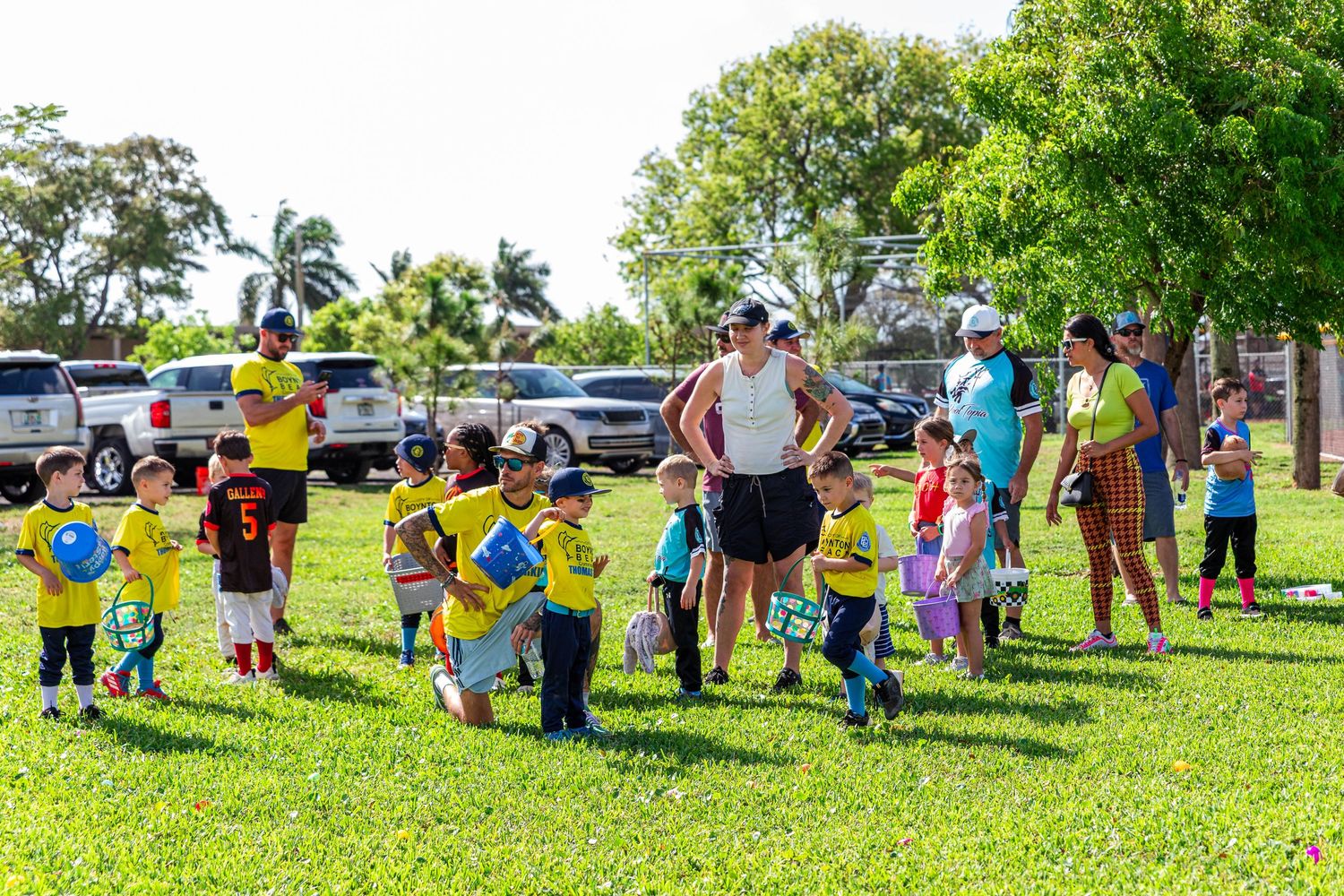 Young children in yellow shirts gather with adults for a kids soccer event on a sunny green park lawn.