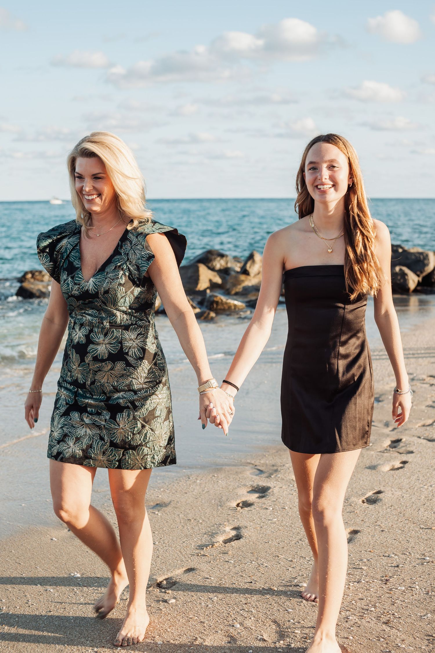 Two people walking together and holding hands on a sunny beach with ocean waves and rocky shore.