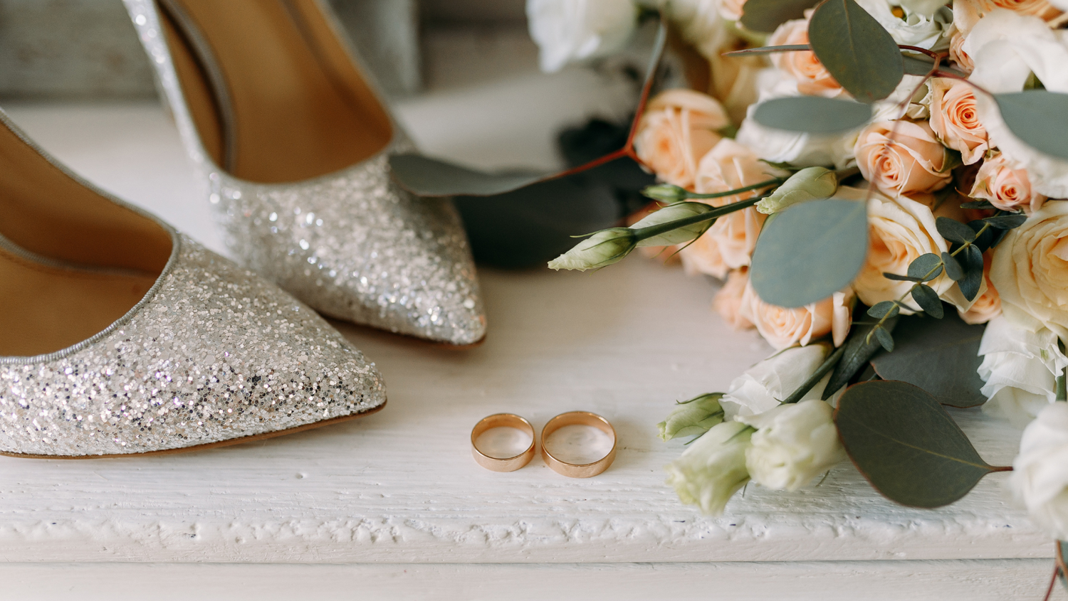 Elegant bridal shoes with glitter detail next to wedding rings and soft pink floral arrangement.