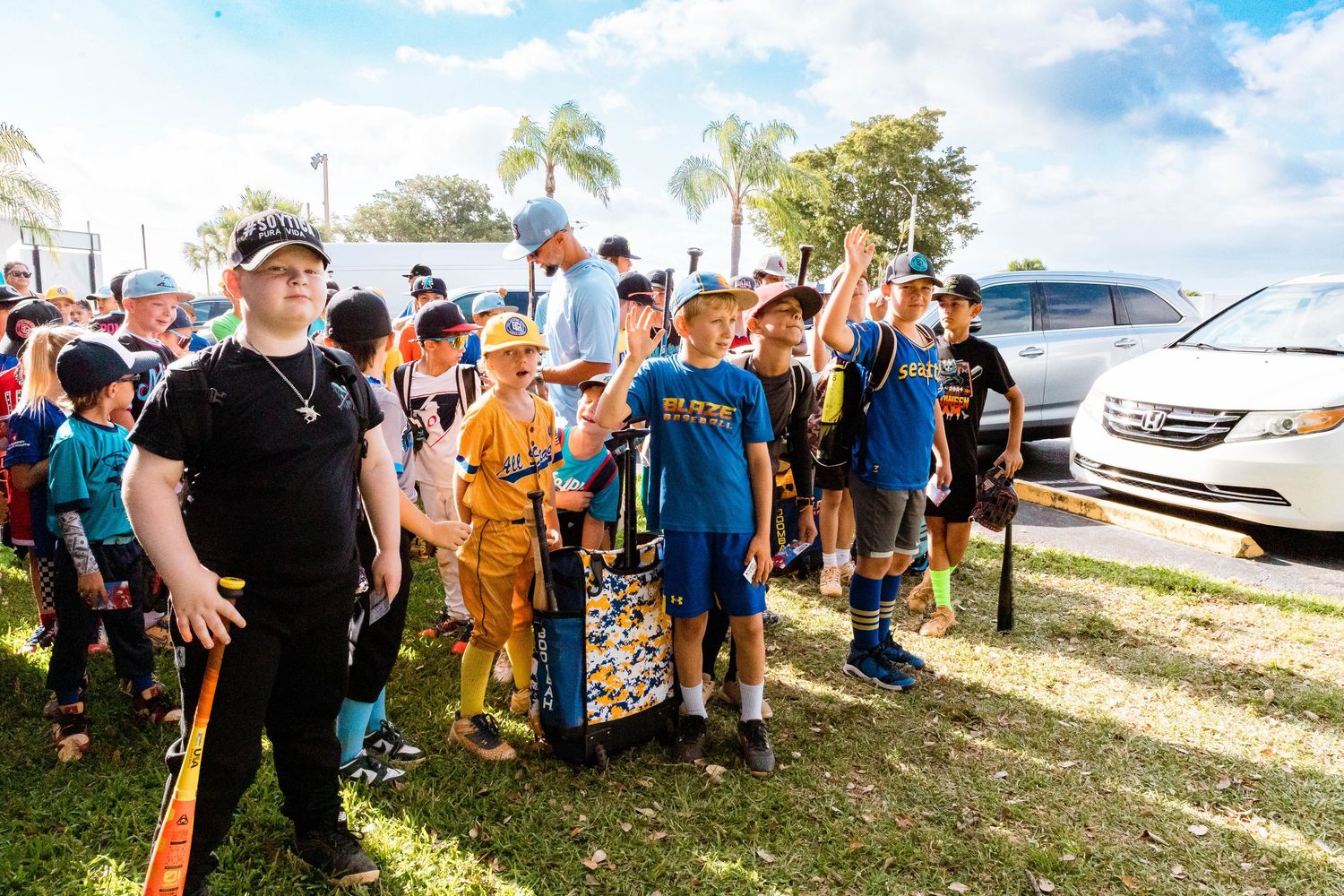 Large group of youth sports players and coaches gather outdoors on sunny day with palm trees and parked cars in background.