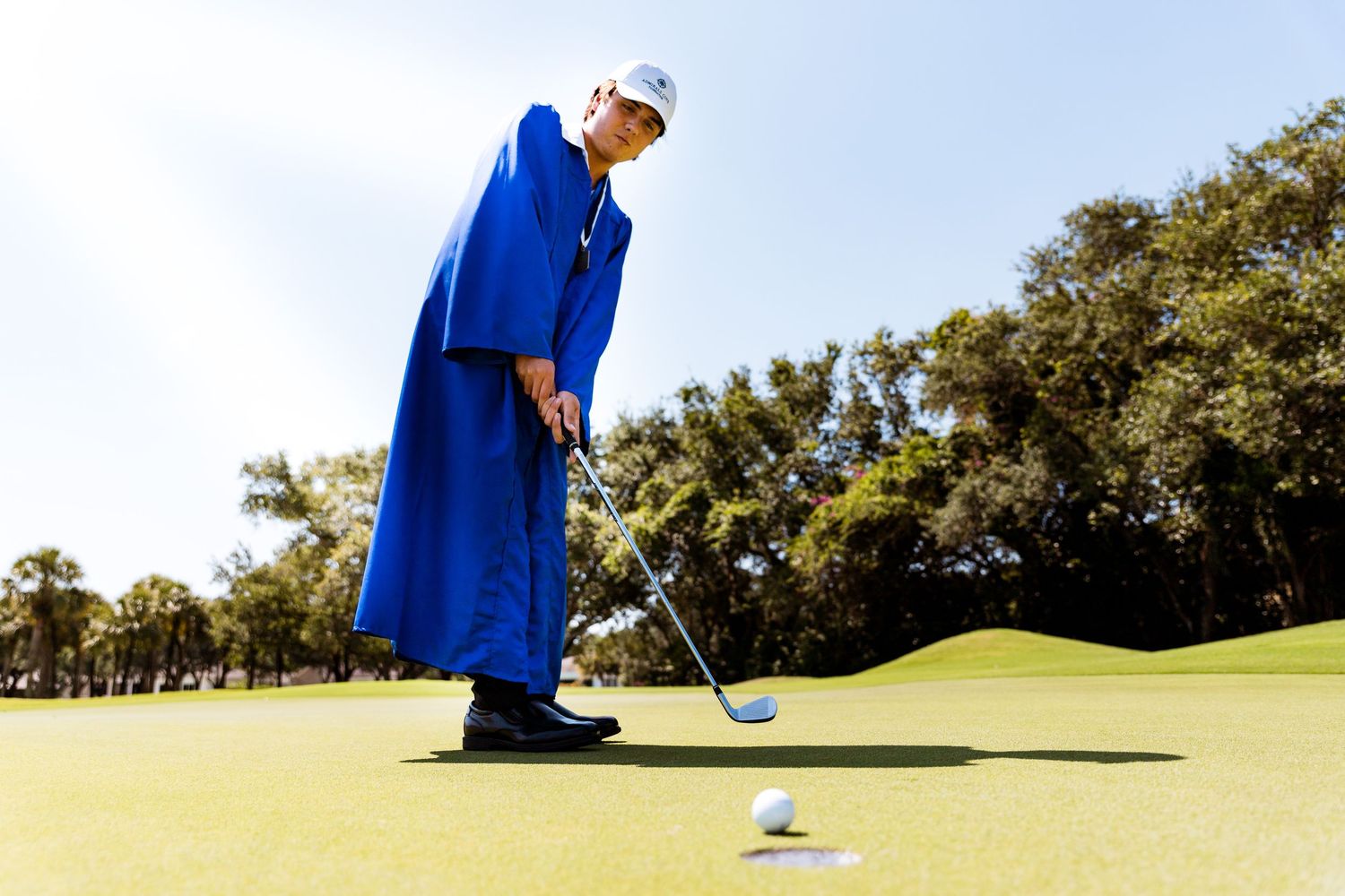 Golfer in flowing royal blue graduation gown attempts putt on sunny green at golf course.