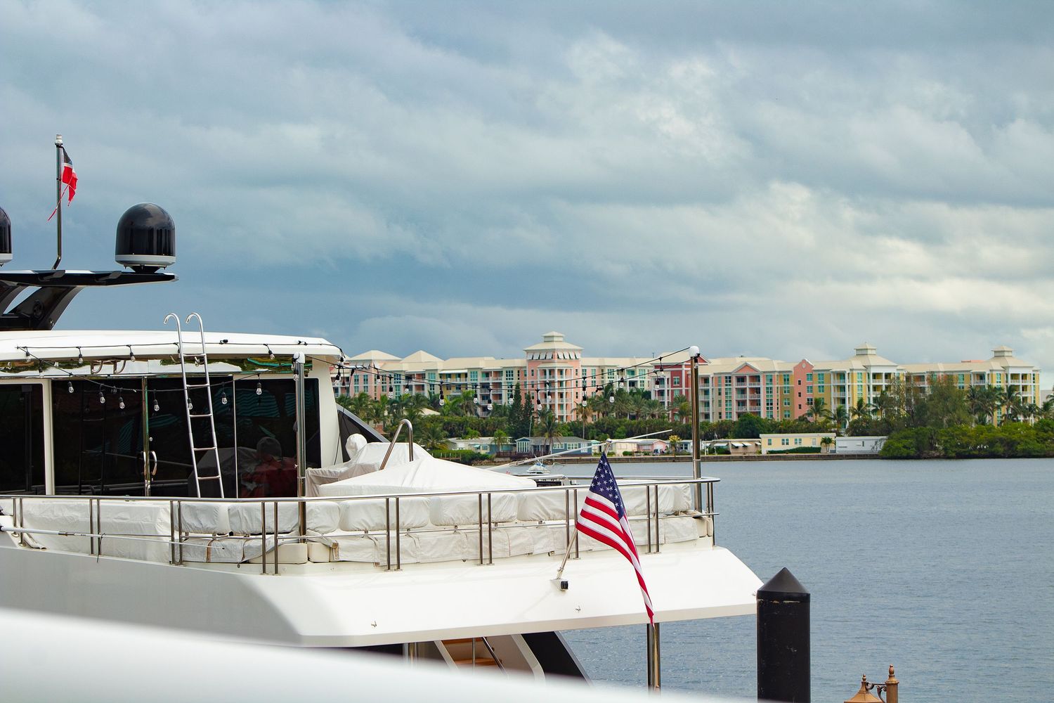 Luxury yacht with American flag floating near waterfront condominiums under cloudy skies.