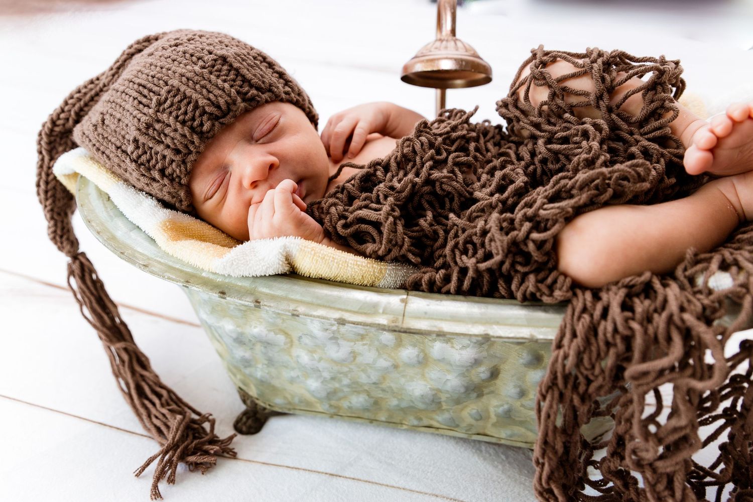 Newborn sleeping in vintage bathtub wearing brown knit hat with textured brown blanket.