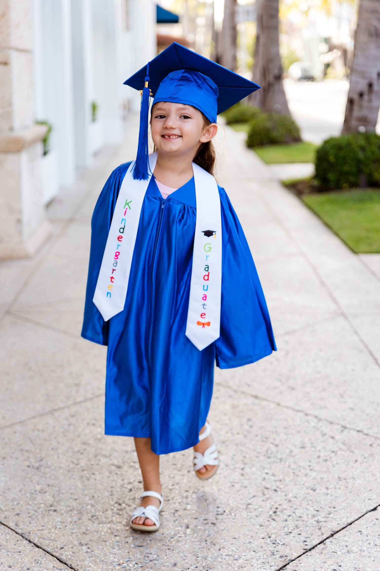 Small graduate in bright blue cap and gown with decorated sash walks smiling on sidewalk wearing white sandals.