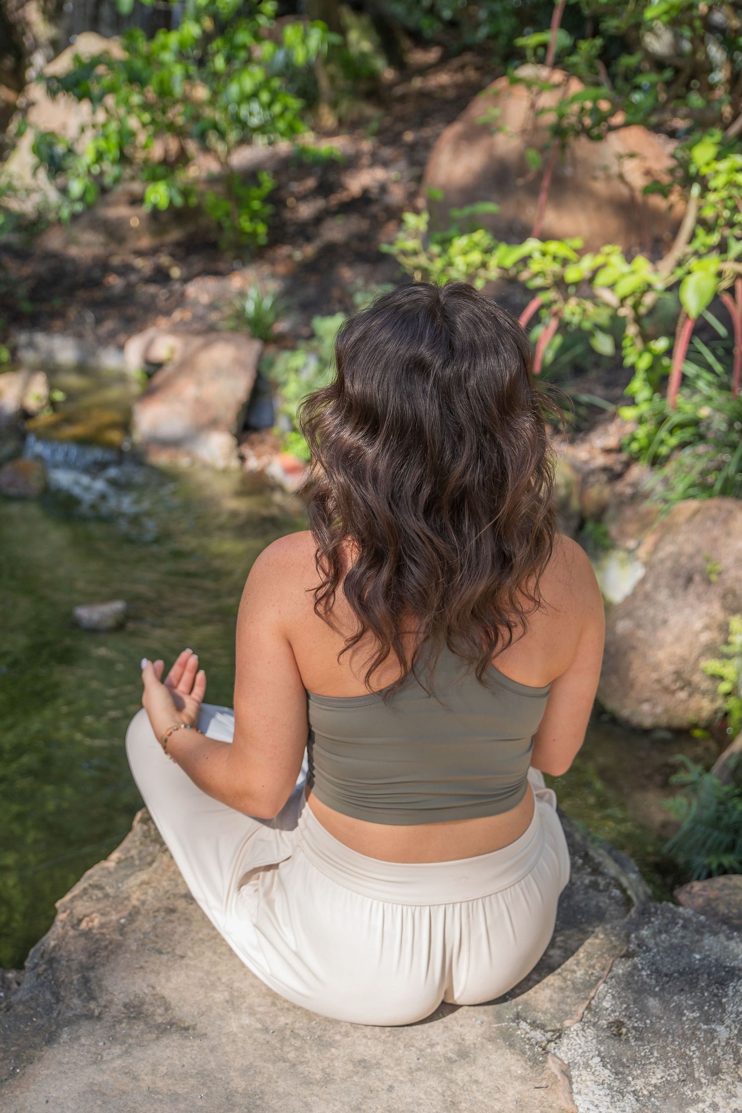 Person in olive tank top and white pants sits peacefully on rocky ledge beside stream in nature setting.