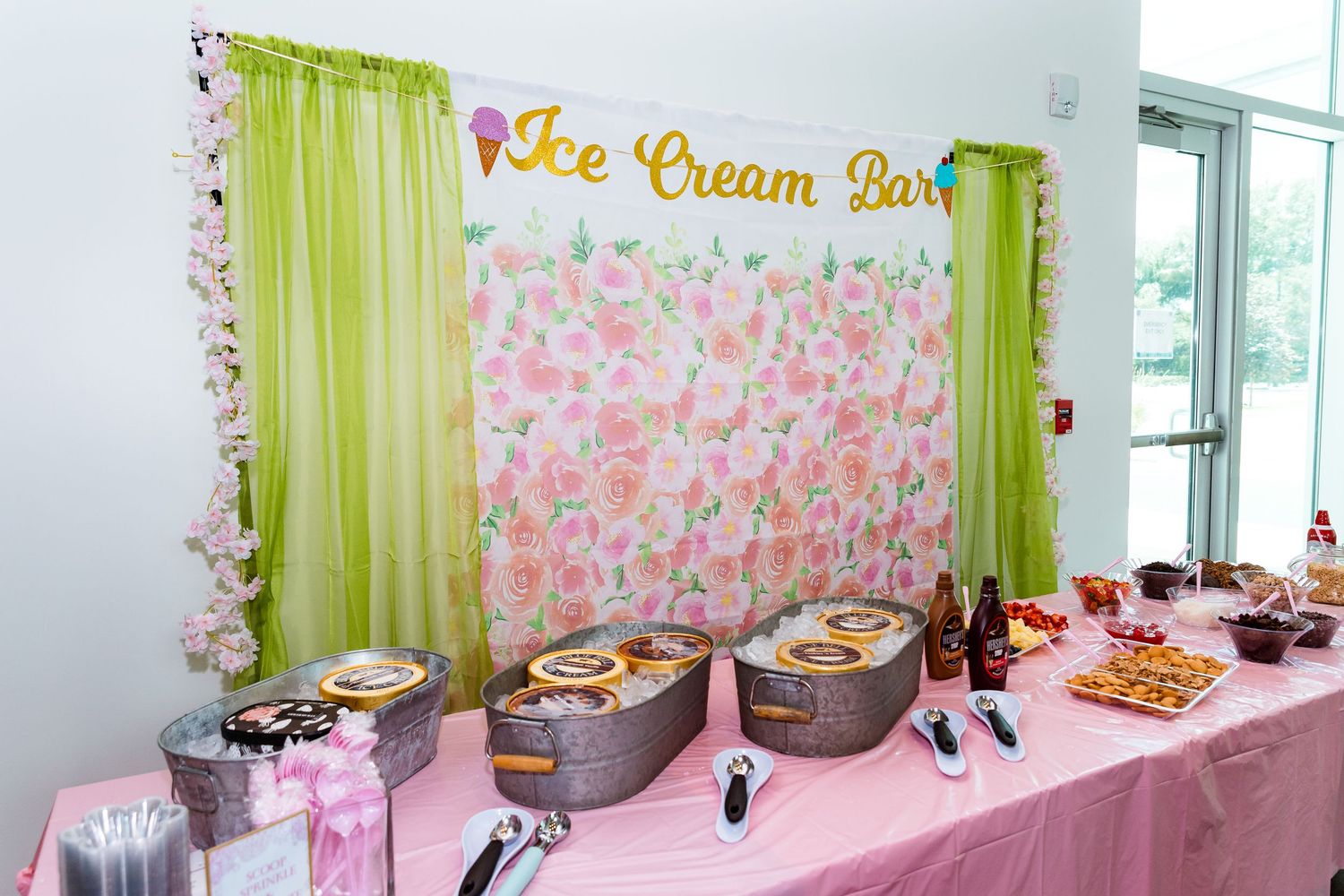 Ice cream bar dessert station with floral backdrop and green curtains displays toppings and treats on a pink tablecloth.