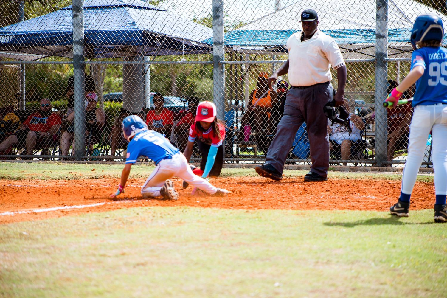 Baseball player slides into base as umpire watches during competitive game on dirt field.