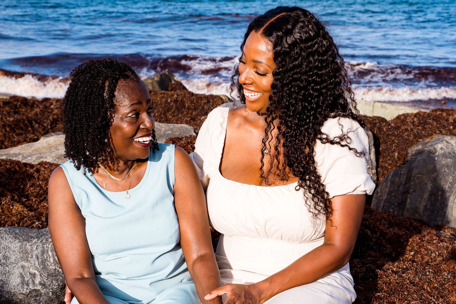 Two people share joyful laughter while sitting together at a rocky beach with blue ocean waves in background.