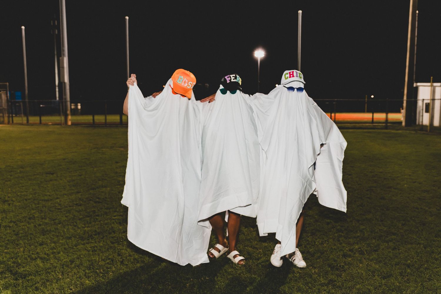 Group in white sheet costumes gathered on grass field at night with stadium lights visible.