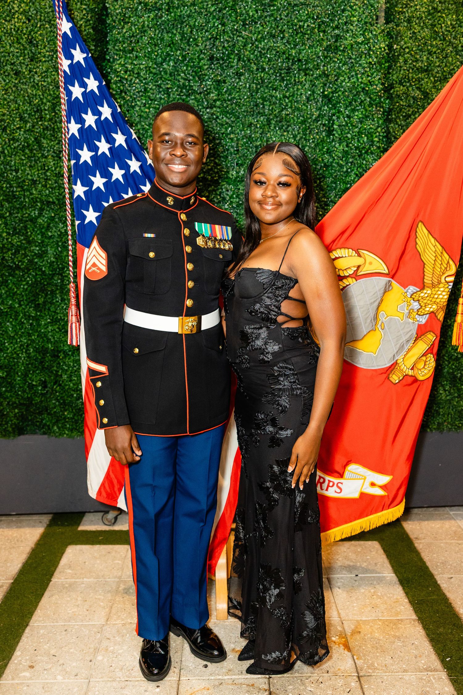 Marine in dress blues uniform poses with companion in formal black gown against green hedge backdrop with flags.
