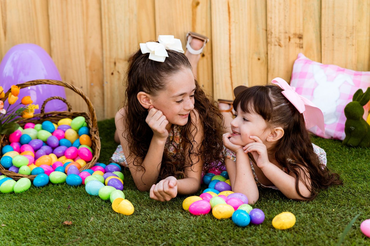 Two people in matching dresses lying on grass with colorful Easter eggs scattered around them.