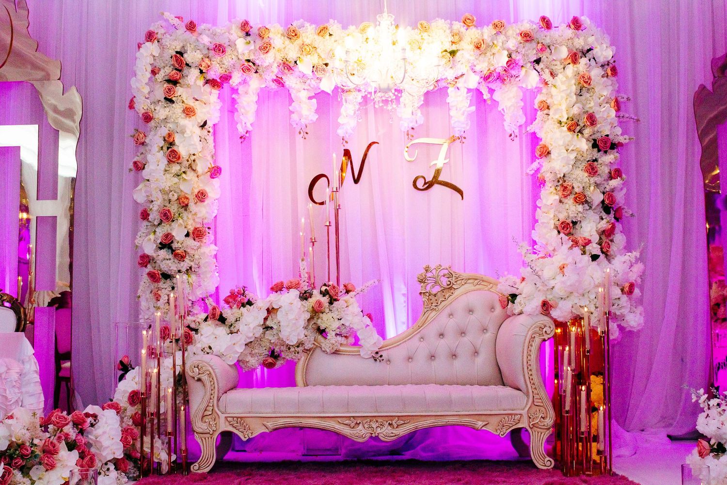 Elegant white loveseat under floral arch with pink lighting and romantic decor.