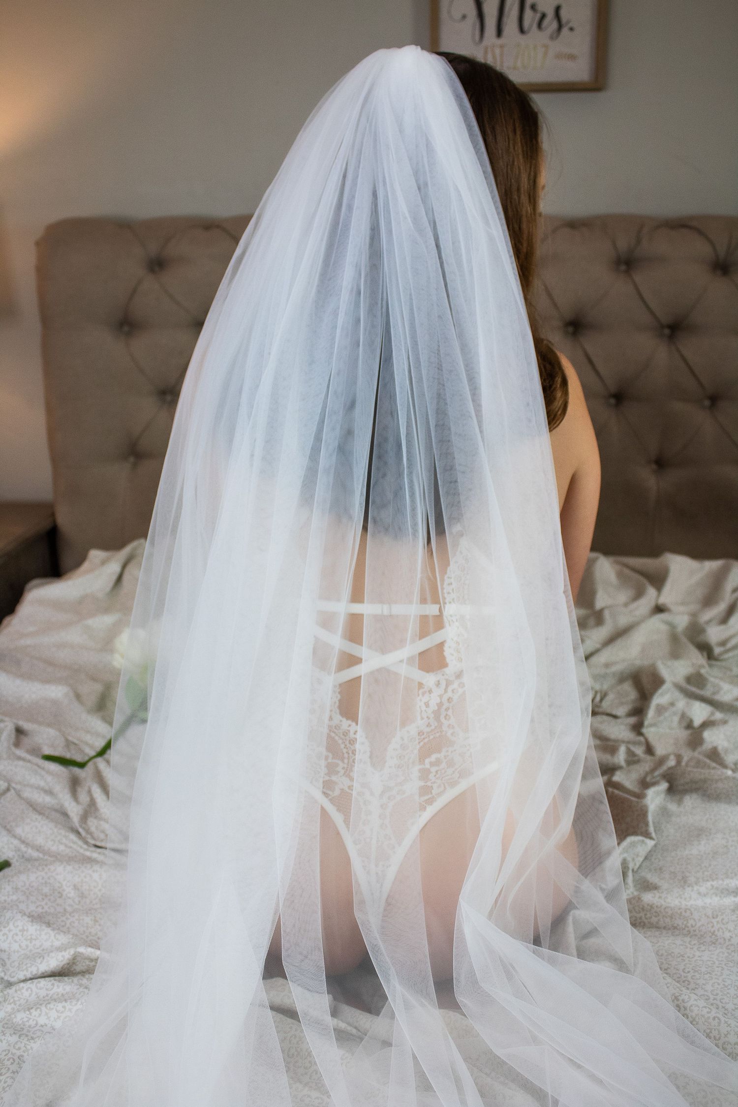 Long flowing wedding veil cascades over white lace lingerie on gray tufted headboard.