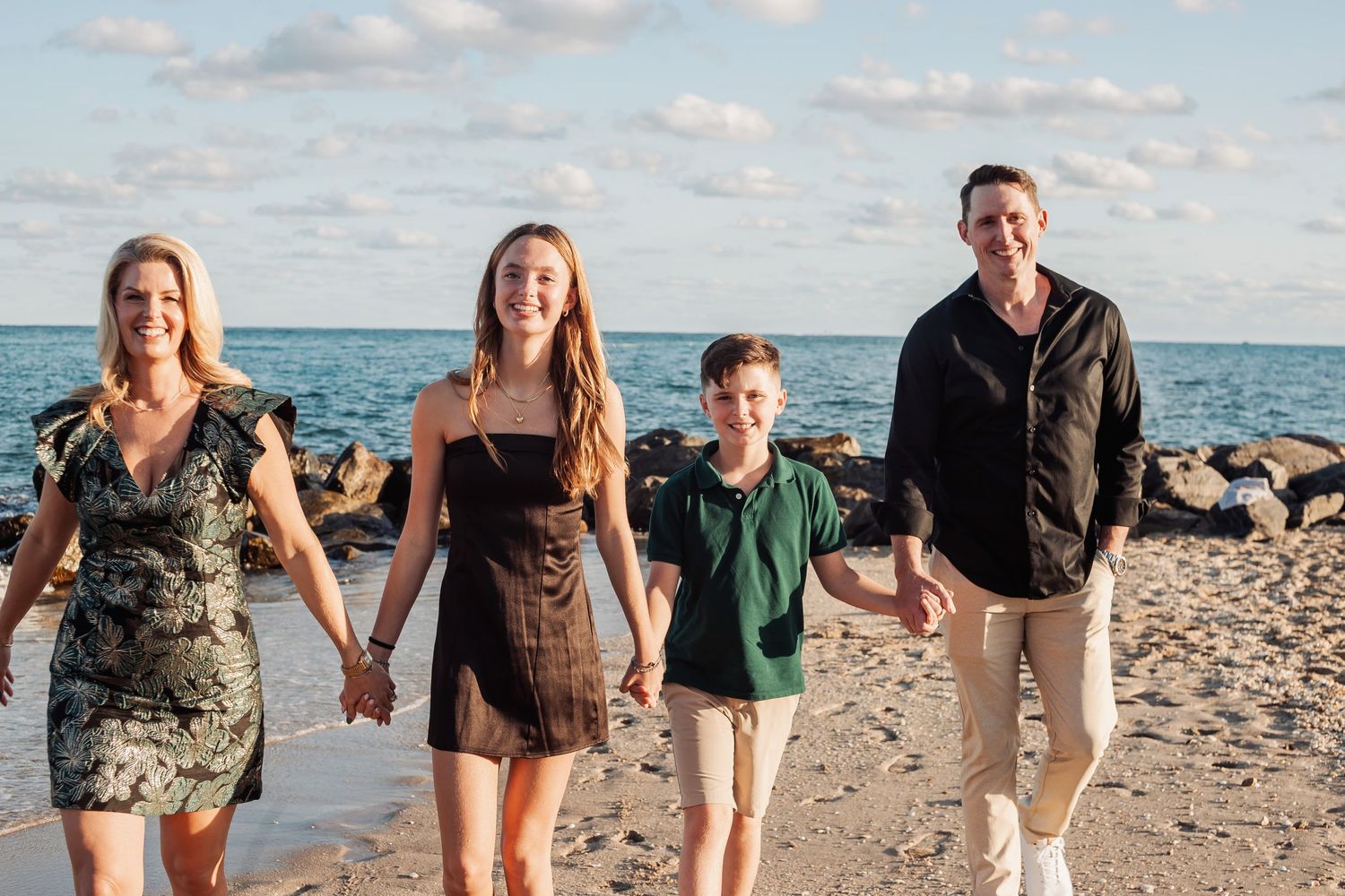 Family holding hands while walking along tropical beach with clear blue waters and rocky shoreline.